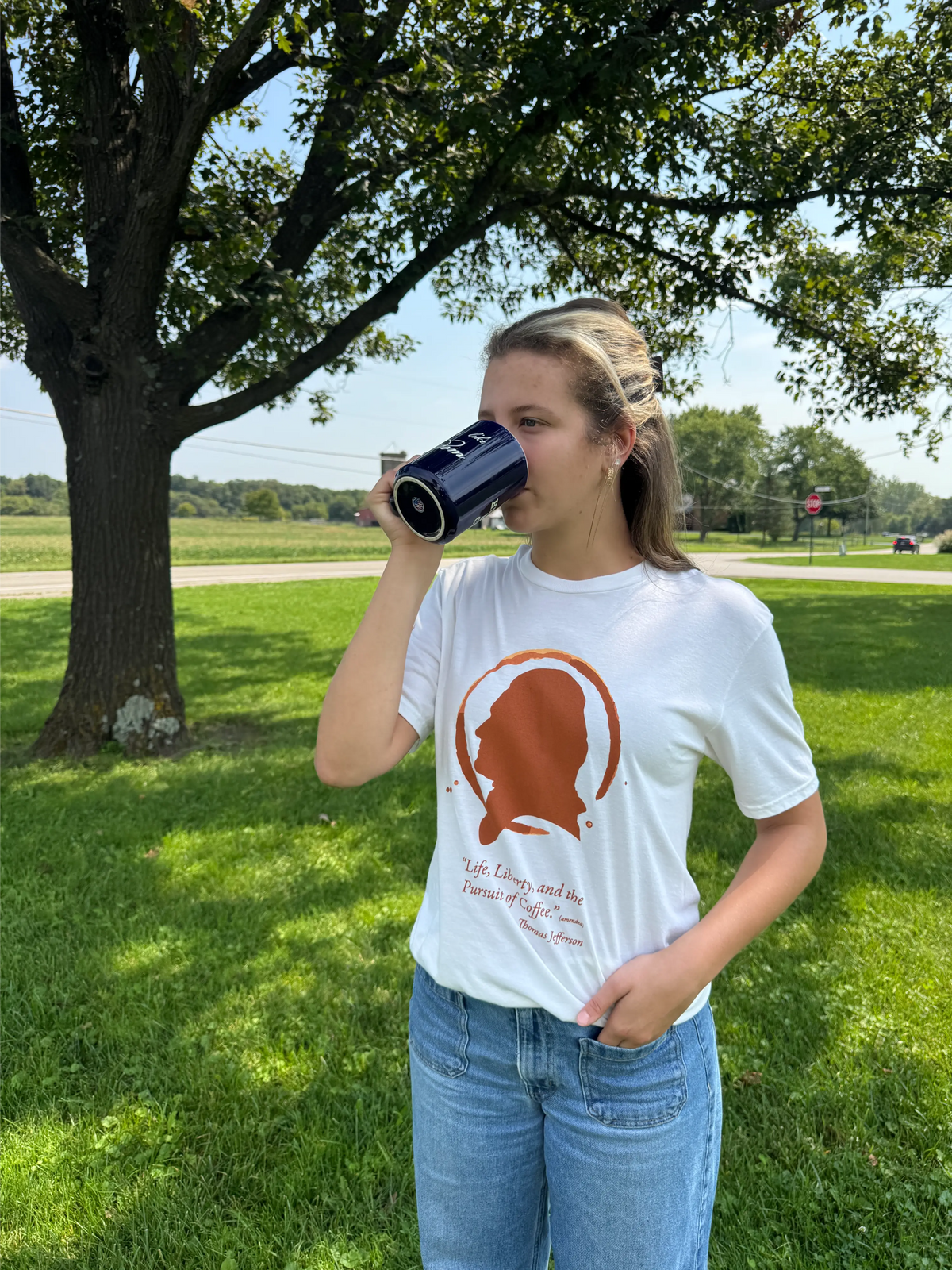 A young woman stands on a grassy lawn under a large tree, drinking from a dark blue can. She is wearing a white graphic t-shirt with a brown silhouette design and blue jeans.